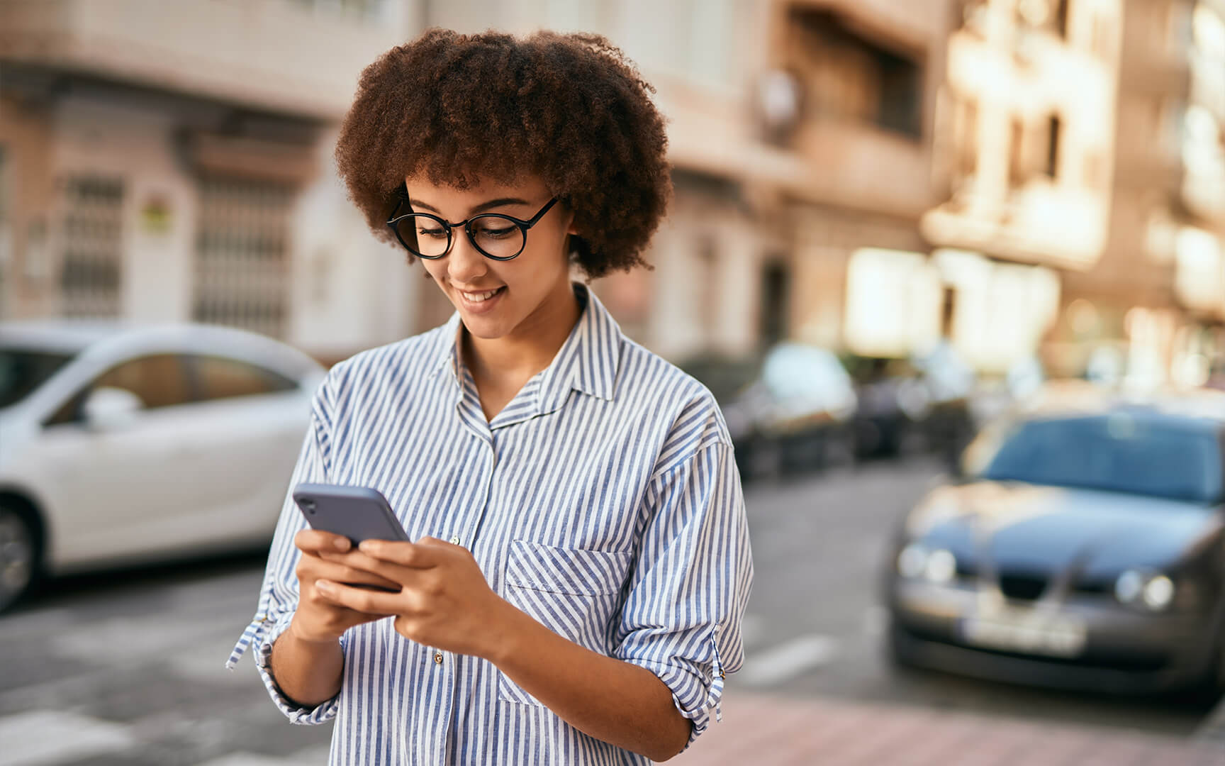 Person standing on a city street, wearing a striped button-down shirt and holding a smartphone, with parked cars and residential buildings in the background.
