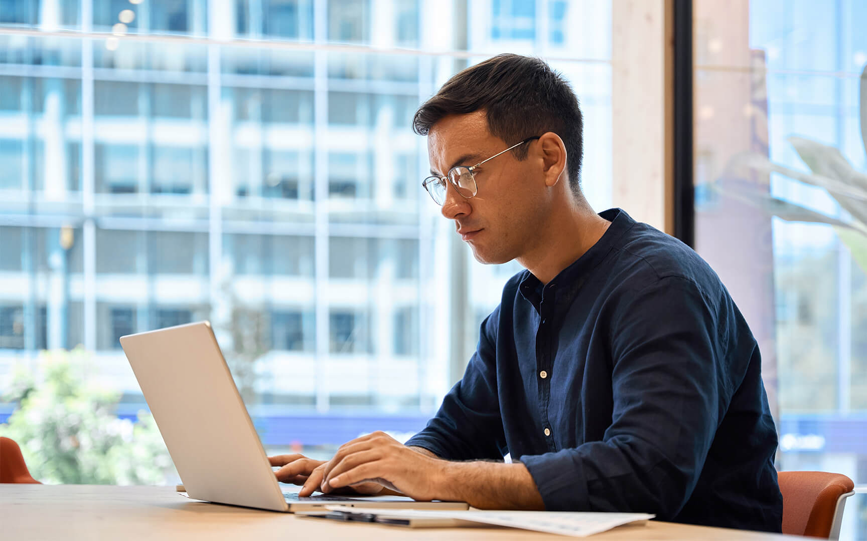 Person working on a laptop at a desk near a large window, with modern office buildings visible outside.