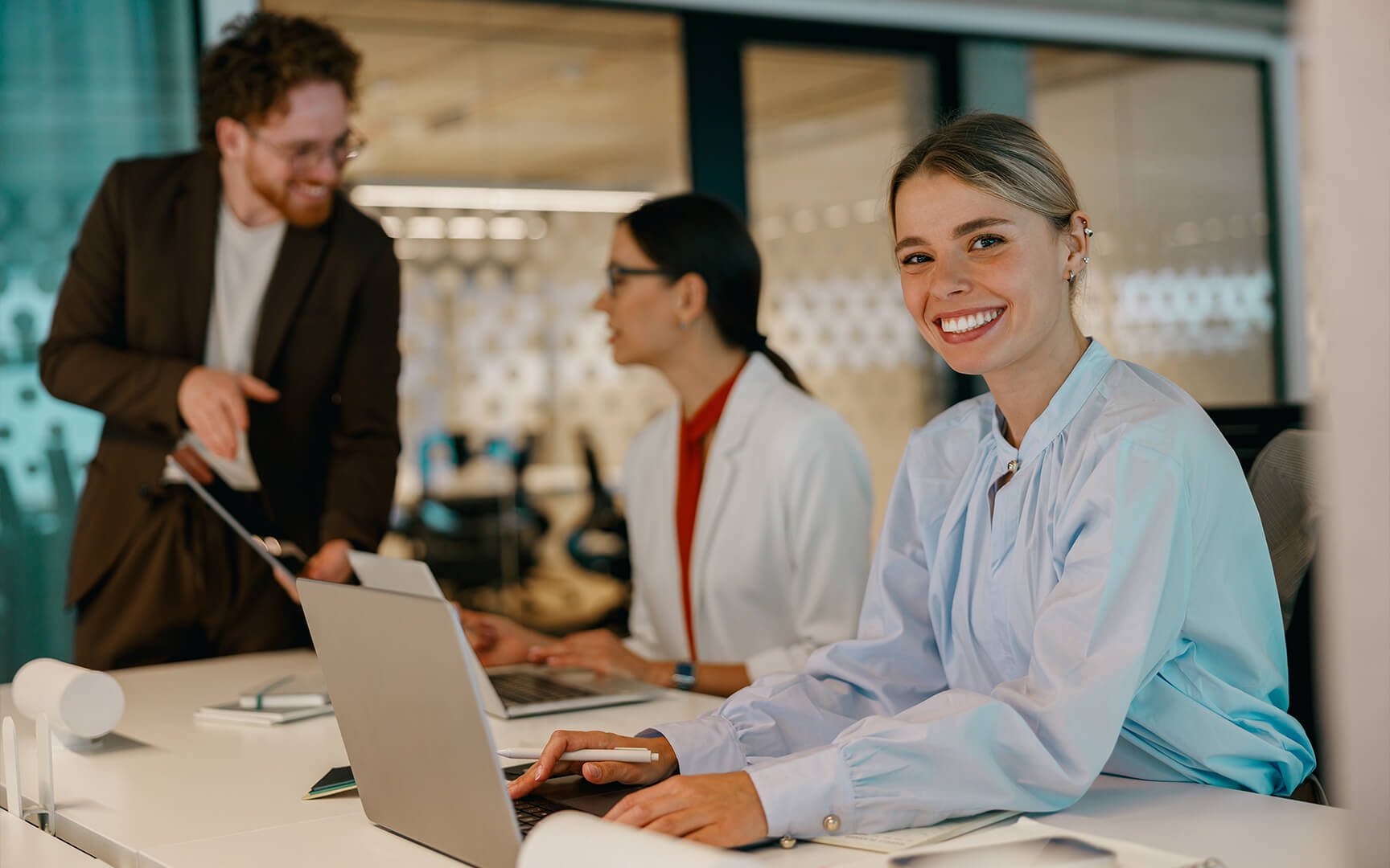 Three people working at laptops in a modern office, with one person in the foreground seated at a desk and two colleagues collaborating in the background.