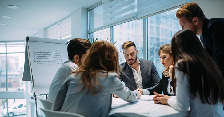A group of professionals seated around a meeting table in a modern office, reviewing documents together, with a flip chart displaying a line graph in the background.