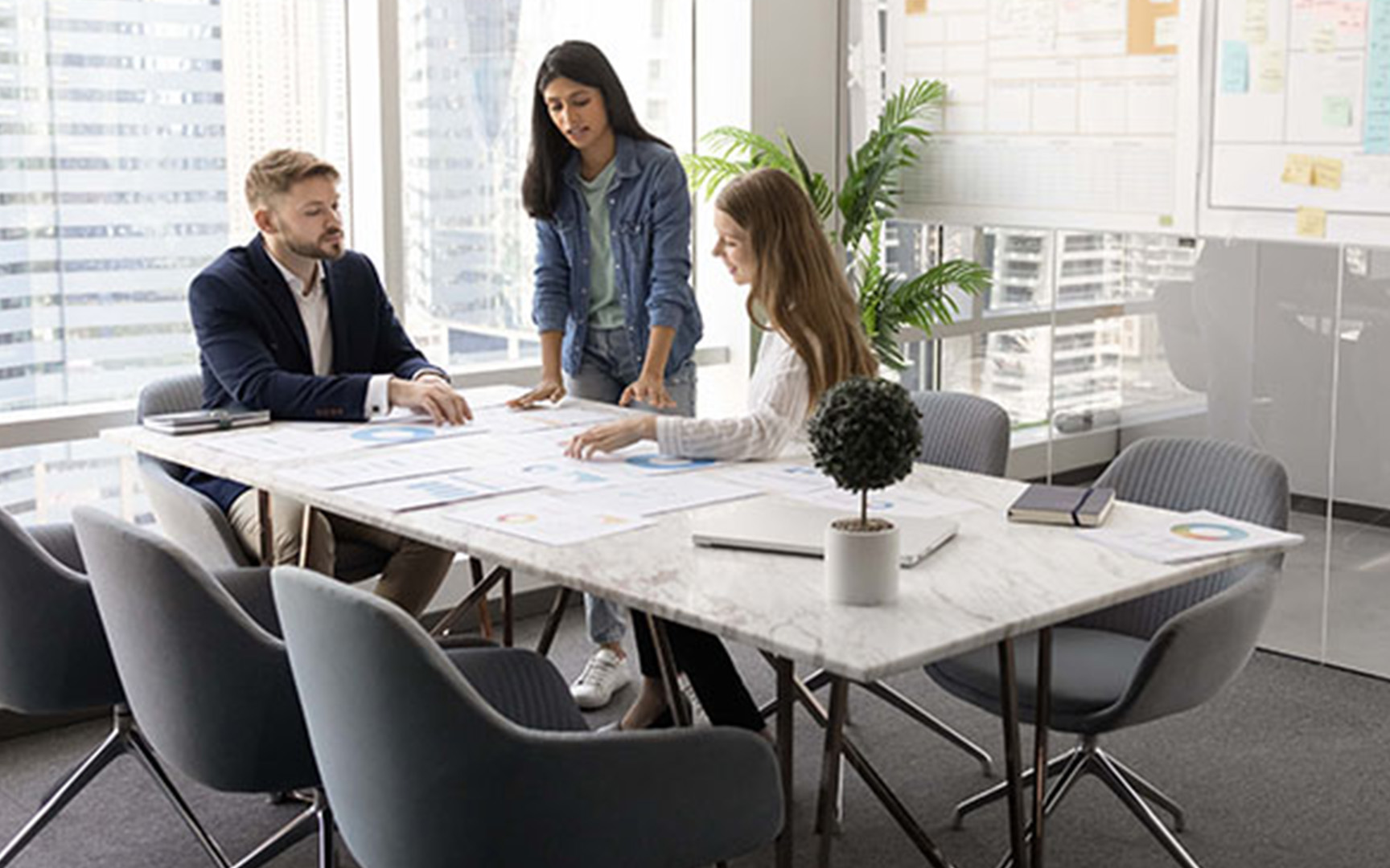 Three professionals collaborating around a meeting table reviewing printed charts and data sheets in a modern office setting, with large windows, sticky notes on a wall, and a structured project planning environment.