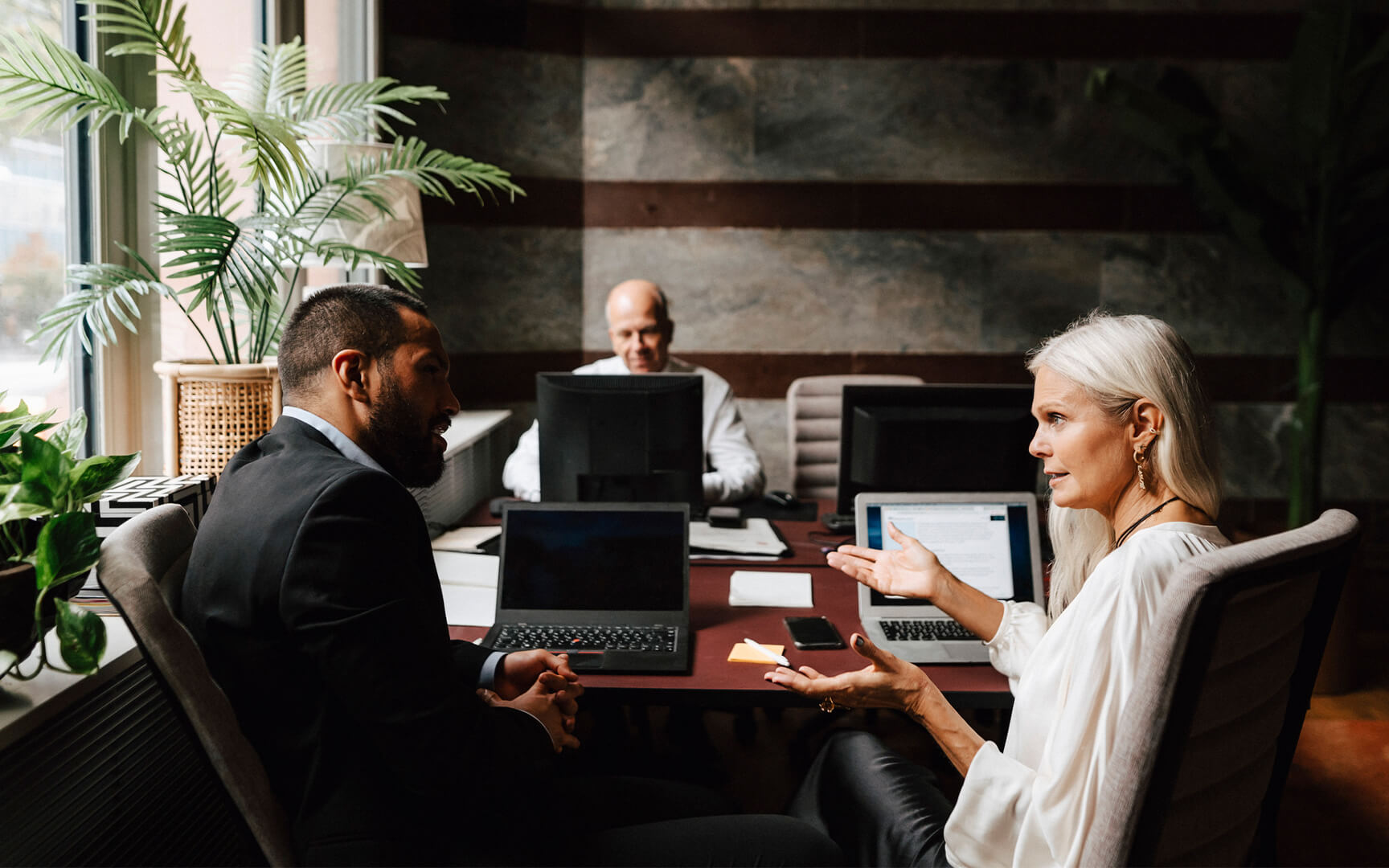 Three professionals sit around a meeting table with laptops, discussing work in a modern office. 