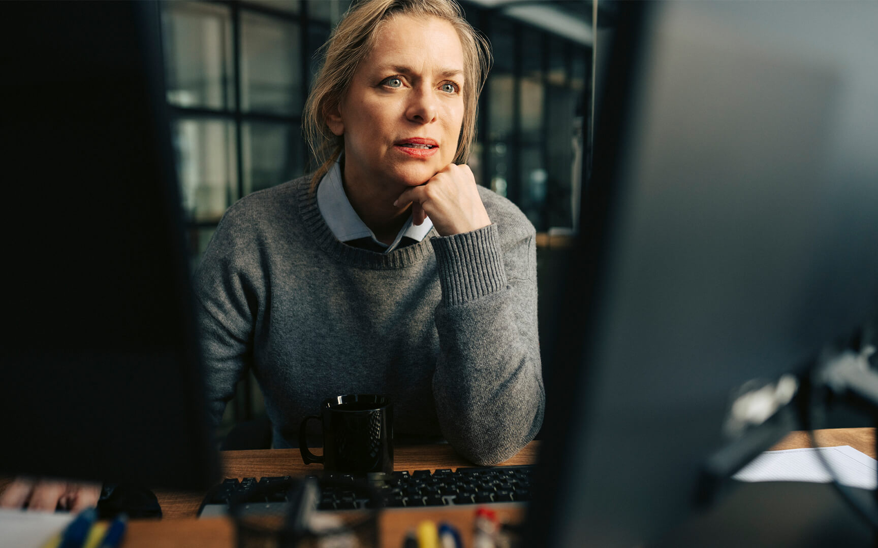 Person seated at a desk looking at a computer screen, with a keyboard, coffee mug, and office items in the foreground.