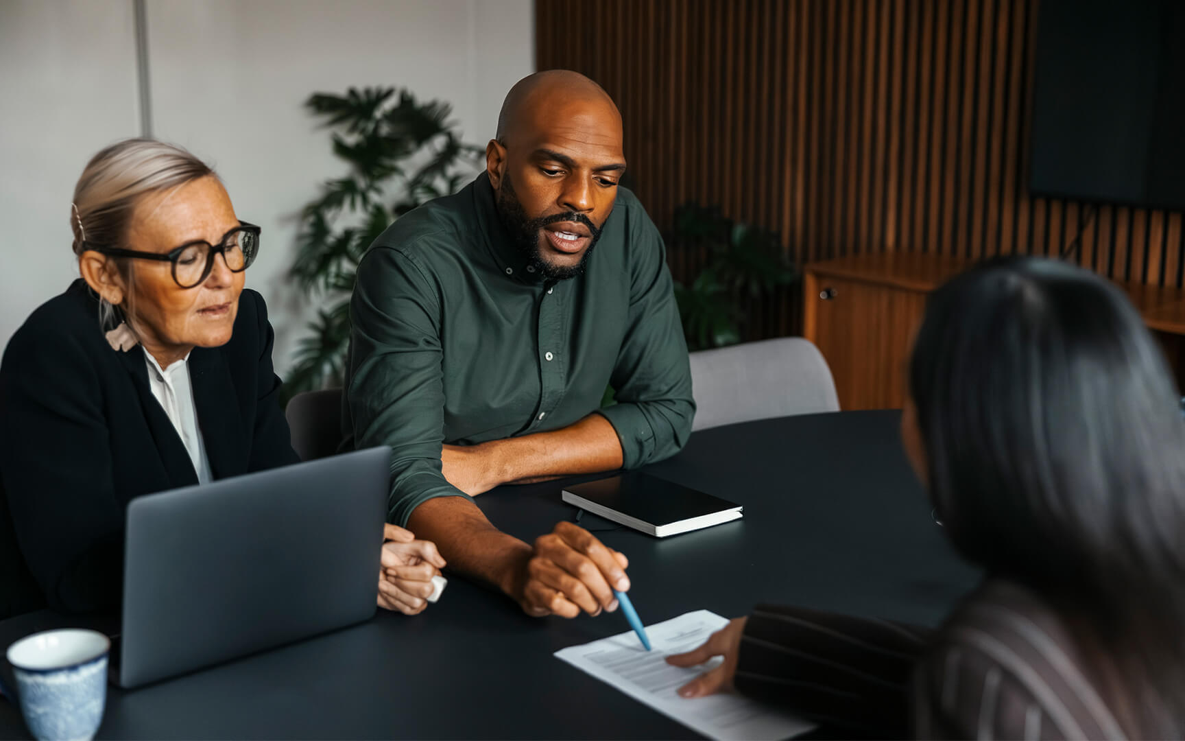 Three people seated at a meeting table reviewing a document together, with one person pointing at the page while another takes notes on a laptop.