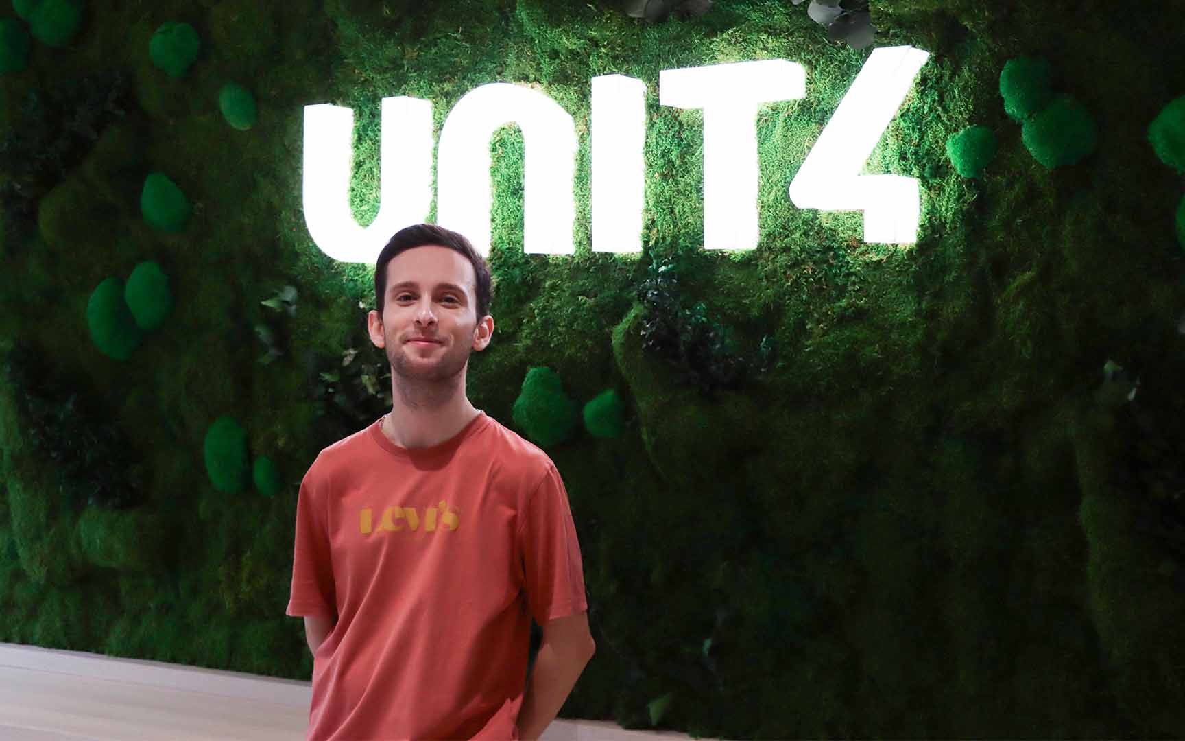 Man standing in front of a green moss wall with a bright illuminated “UNIT4” sign, wearing a red Levi’s T‑shirt in an indoor office setting.