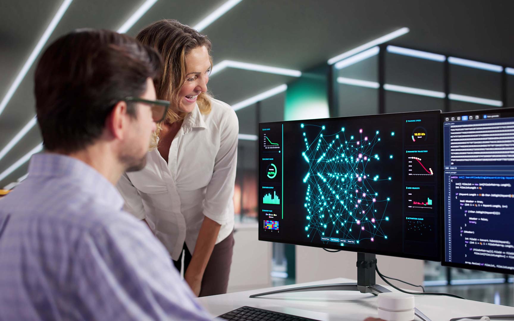 Two professionals collaborating at a desk, reviewing data visualizations and code on a widescreen monitor displaying a network graph and analytics dashboards in a modern office environment.