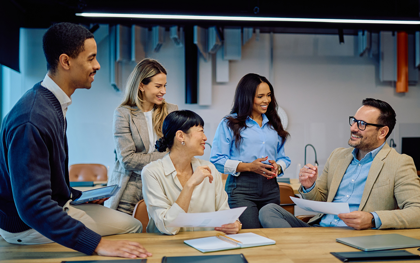 A group of professionals collaborating in a modern office setting, reviewing documents and discussing insights around a table, with laptops and materials visible, highlighting teamwork, planning, and decision-making.