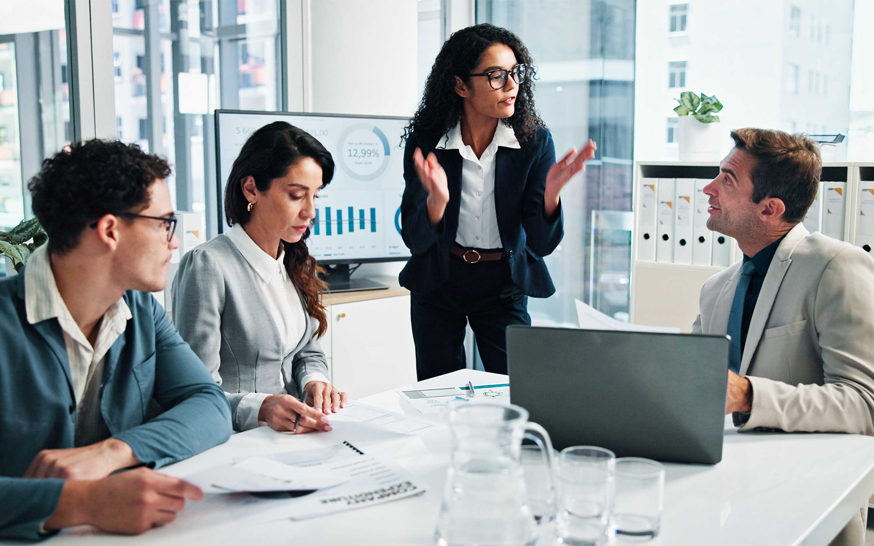 Four professionals in a modern office meeting room sit around a table reviewing printed documents and a laptop, while one person stands and gestures during a discussion. A screen behind shows charts and data, with binders and glassware visible on the table.