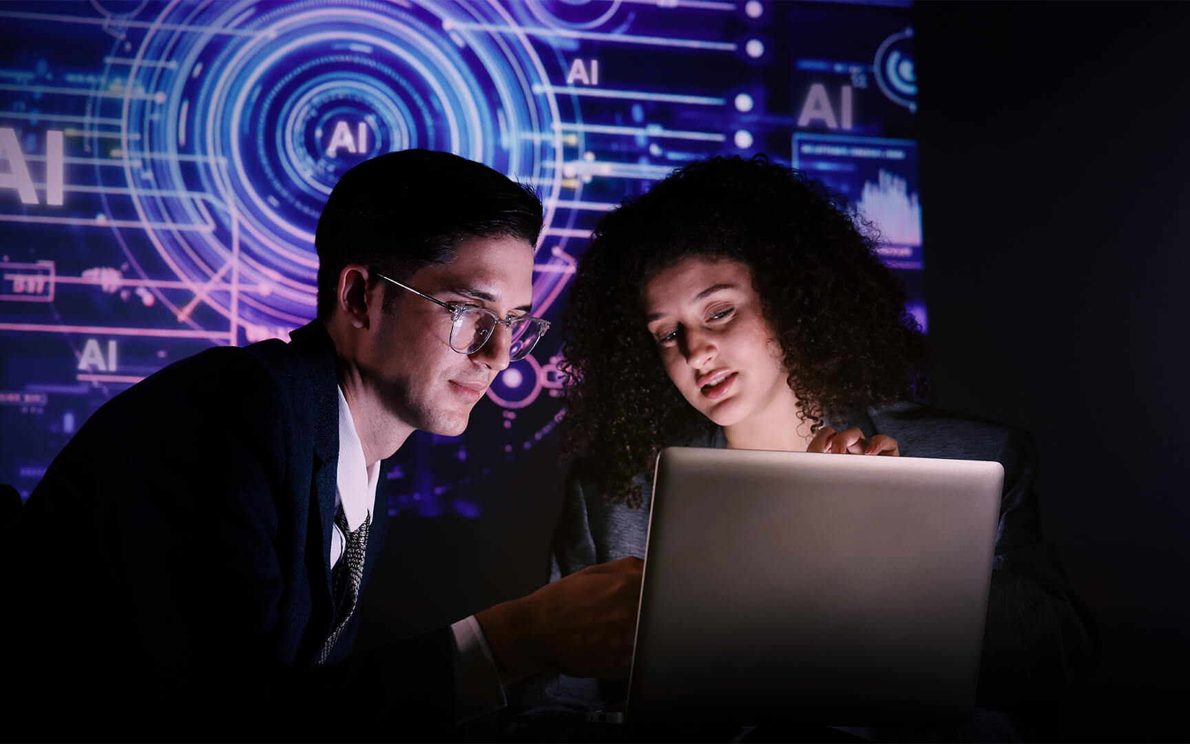 Two professionals working together on a laptop in a dark office setting, with digital AI interface graphics and data visualizations displayed in the background, highlighting the use of artificial intelligence and analytics.