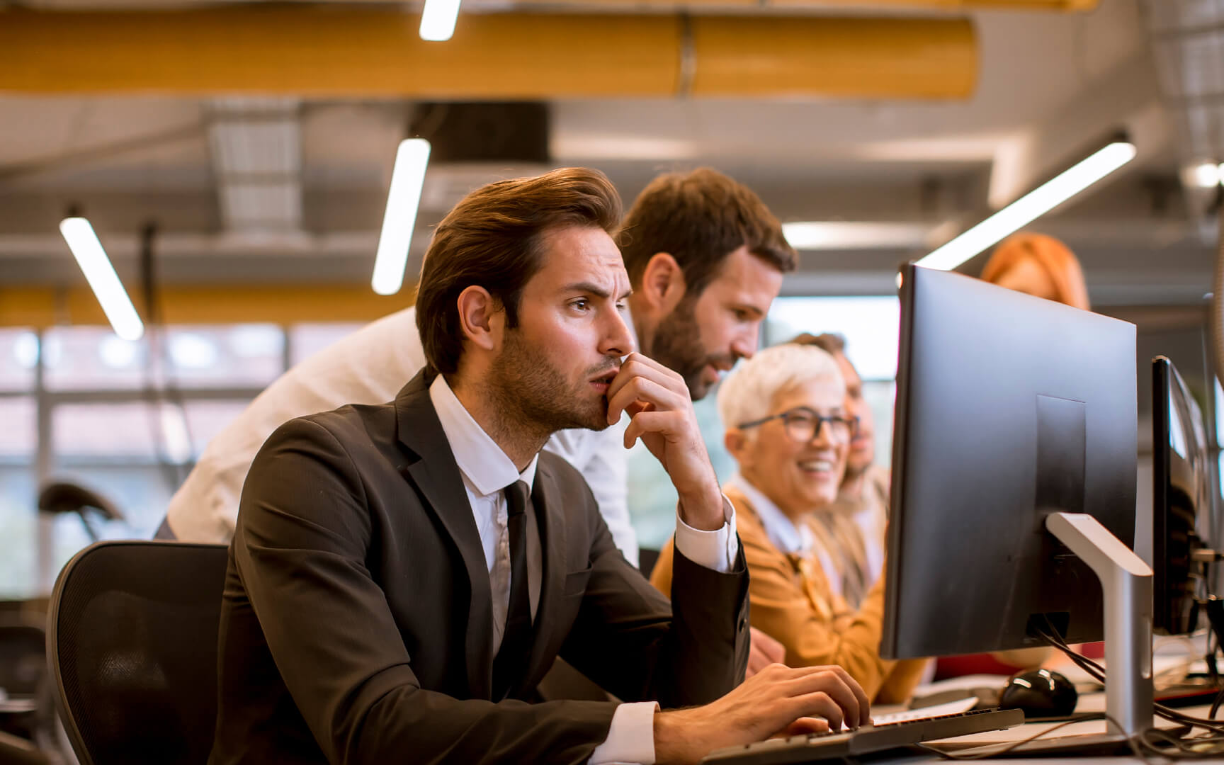 Professional office setting with a group of employees working at desktop computers; a person in the foreground focuses on a screen while colleagues collaborate behind, in a modern workspace with warm lighting.
