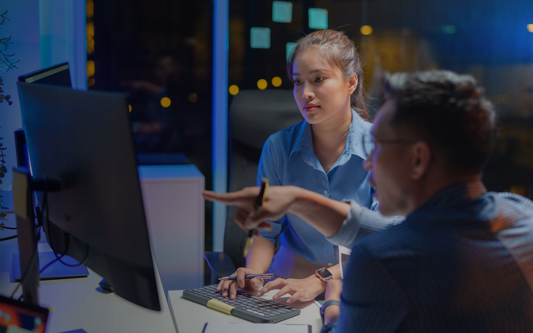 Two colleagues working together at a computer in an office at night, with one pointing at the screen while the other types on a keyboard.