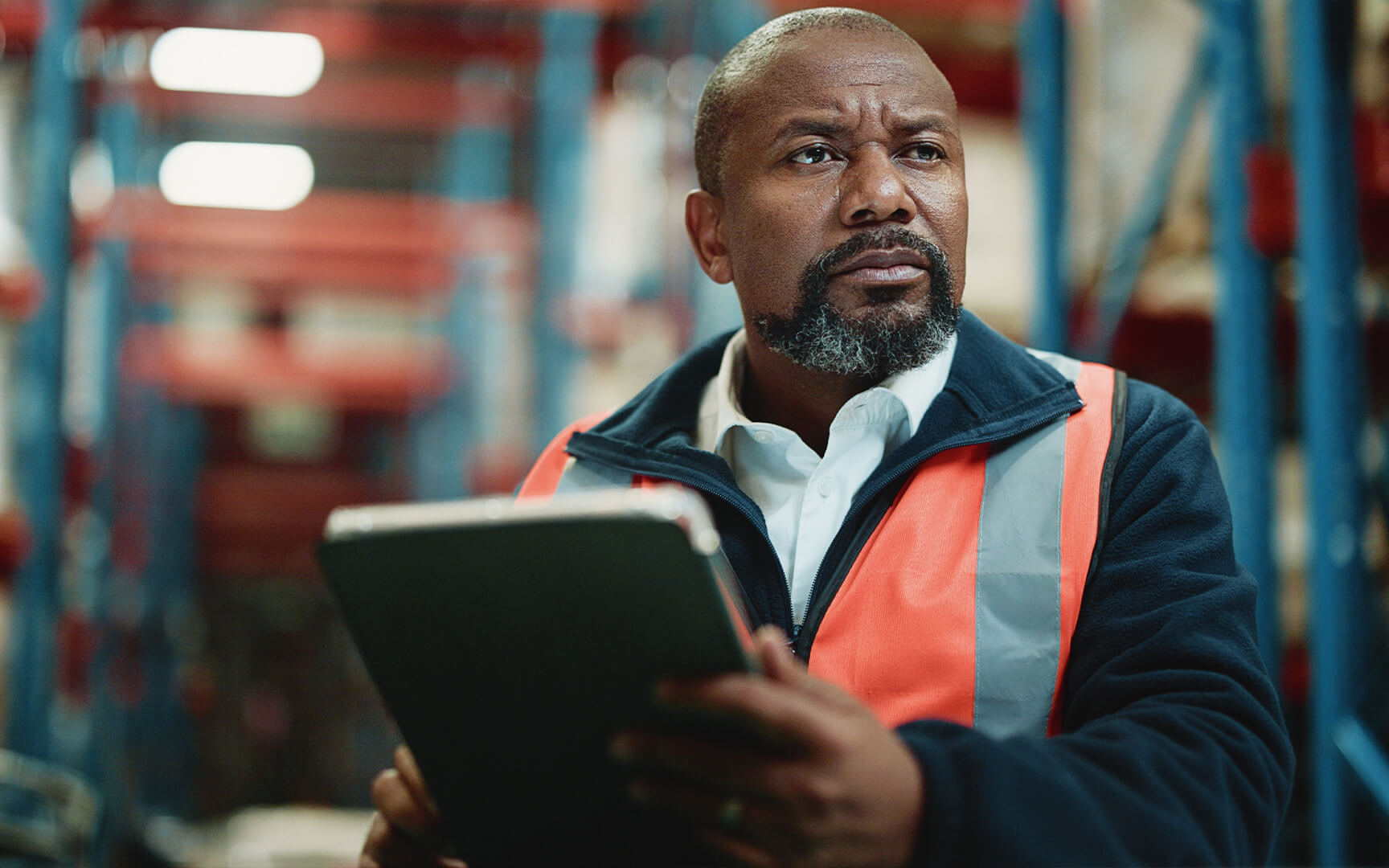Worker wearing a high-visibility orange safety vest holding a tablet in a warehouse or industrial setting, reviewing data or inventory.