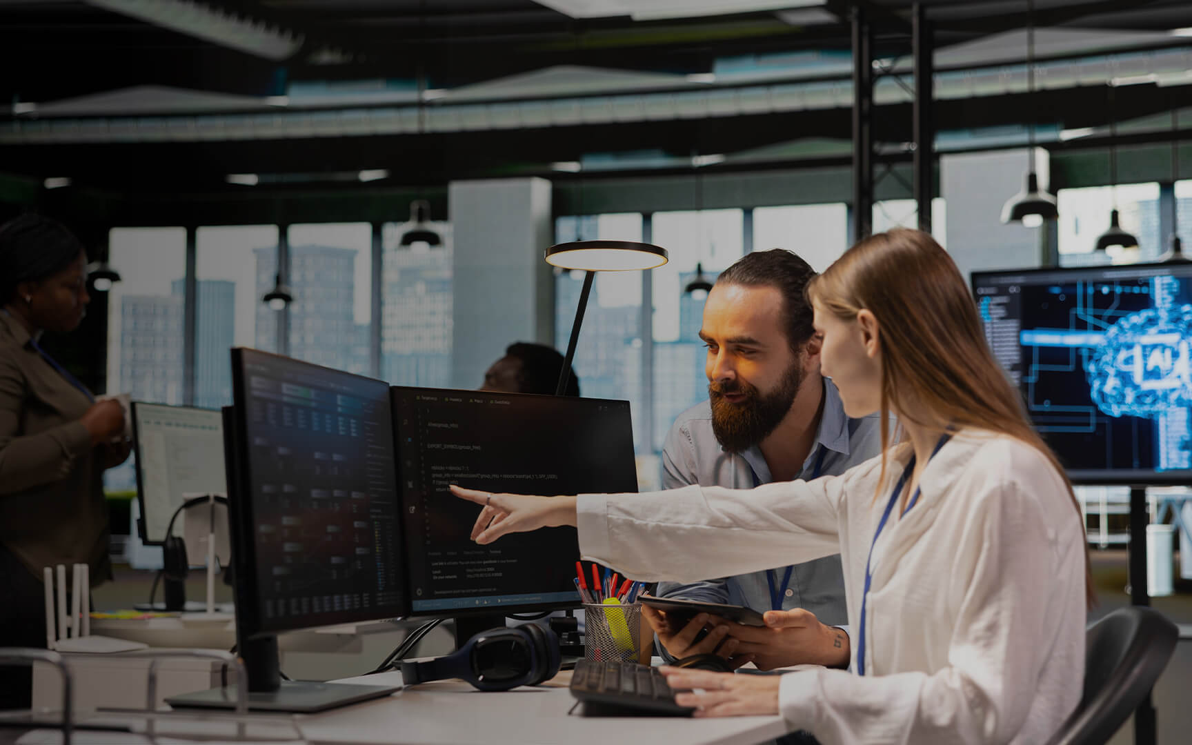Two professionals collaborating at desks in a modern office, reviewing data and code on multiple monitors, with the foreground screen displaying dashboards and analytics, and a cityscape visible through large windows in the background.
