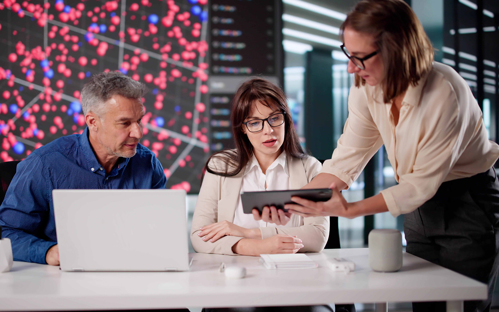 Three professionals collaborate at a desk in a modern office, reviewing information on a tablet and laptop.
