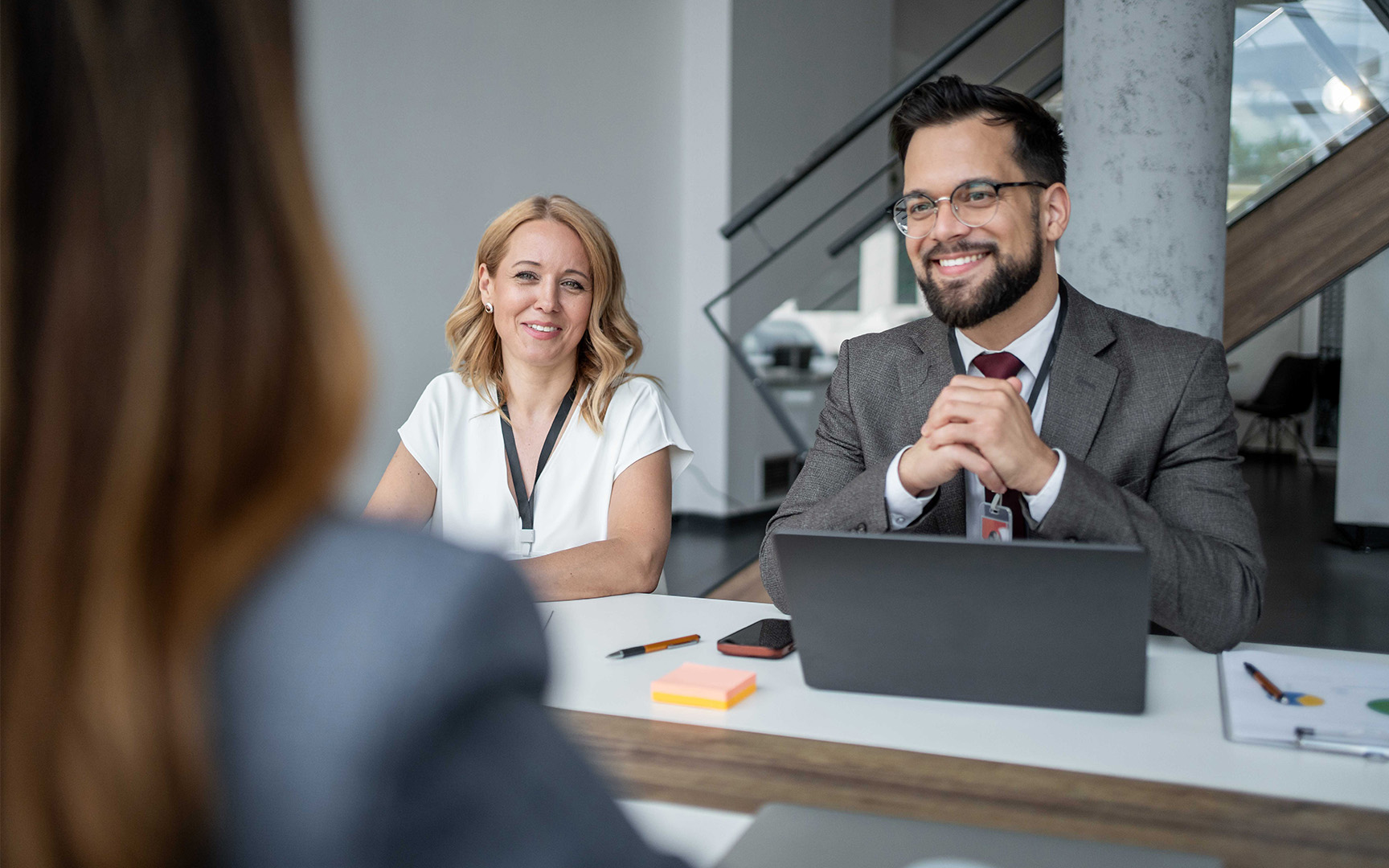 Two professionals sit across a table in a modern office meeting space, facing a person in the foreground.