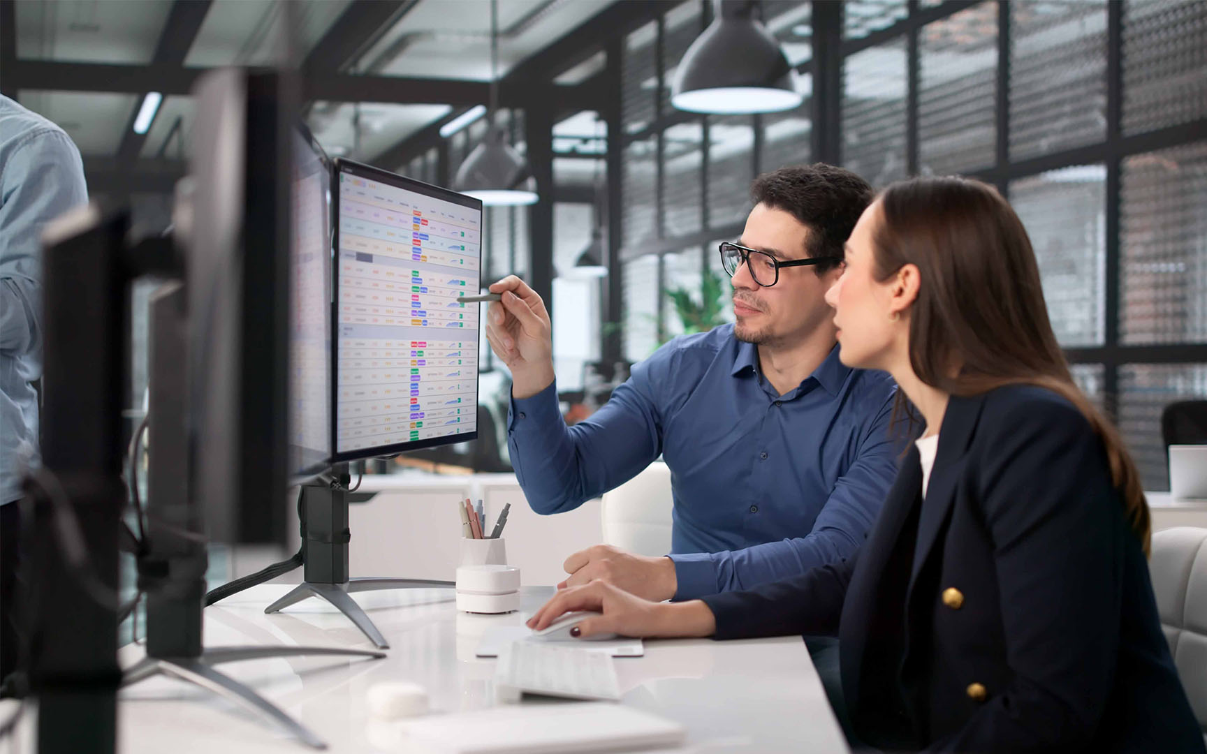 Two professionals sit at a desk in a modern office, reviewing financial or operational data displayed on a computer monitor.