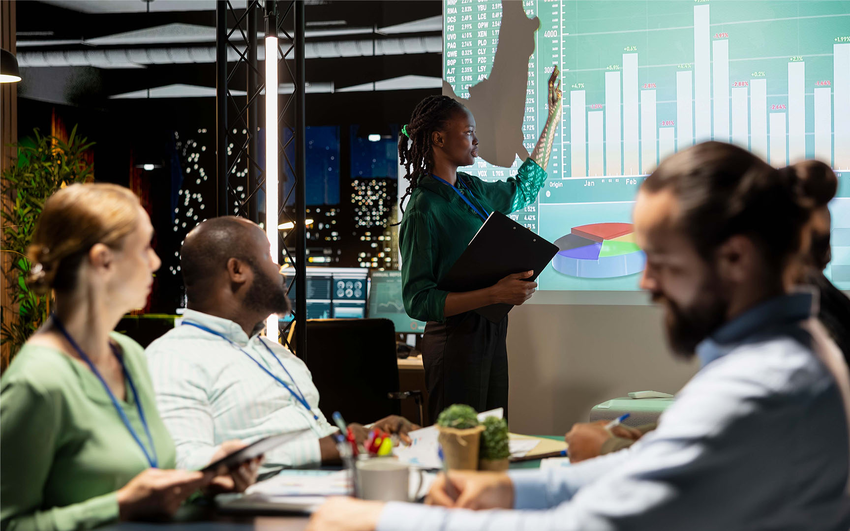 Person presenting workforce analytics and performance dashboards on a large screen during a business meeting, while colleagues review data and reports around a conference table.