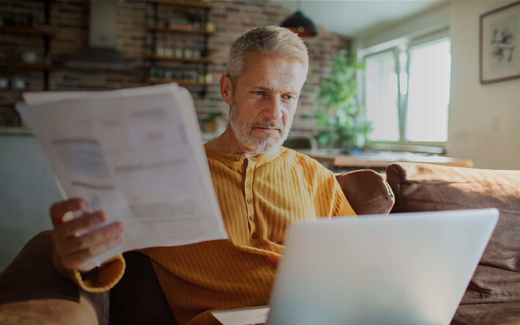 Person seated on a sofa at home reviewing a printed financial document while working on a laptop.