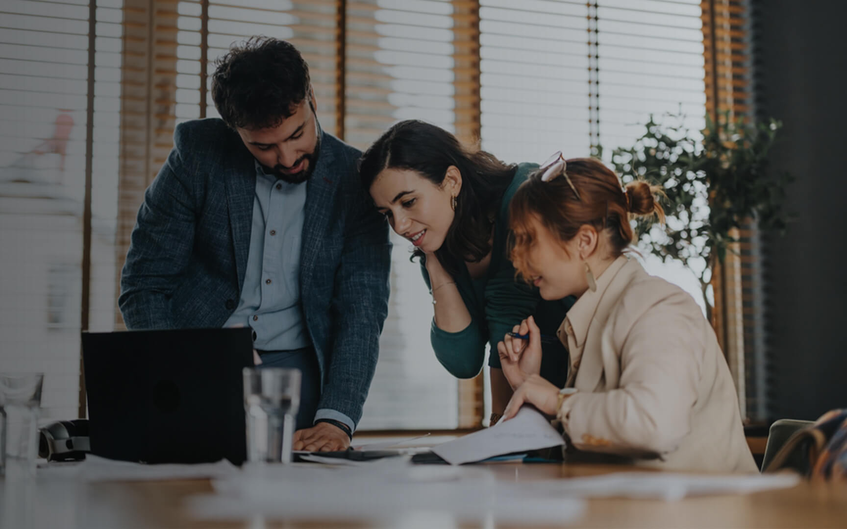 Three colleagues gathered around a laptop and printed documents in a modern office, reviewing procurement data and discussing business decisions during a collaborative planning session.