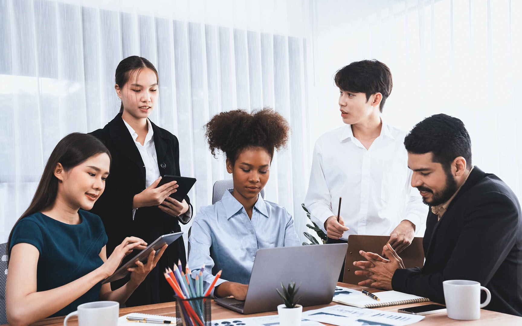Team collaborating around a laptop during a procurement planning meeting, with participants reviewing documents, using tablets, and discussing workflow processes in a modern office environment.
