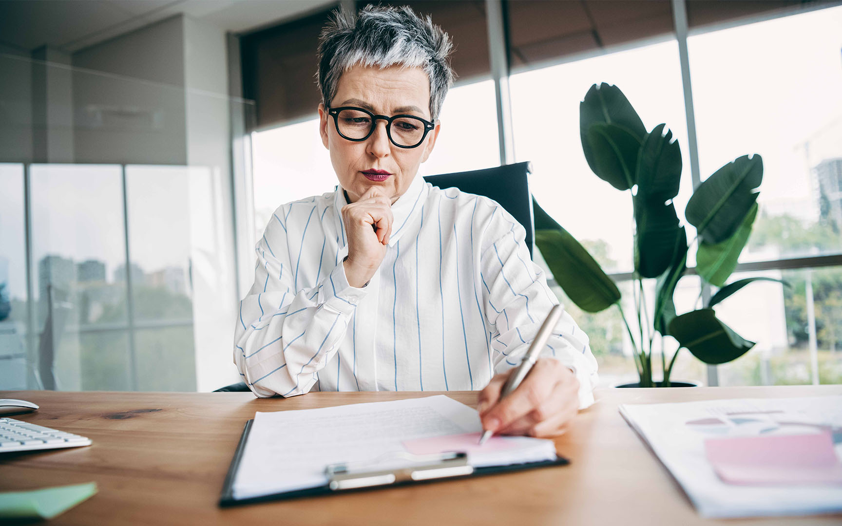 Professional seated at a desk in a modern office, reviewing documents and making notes on a clipboard.