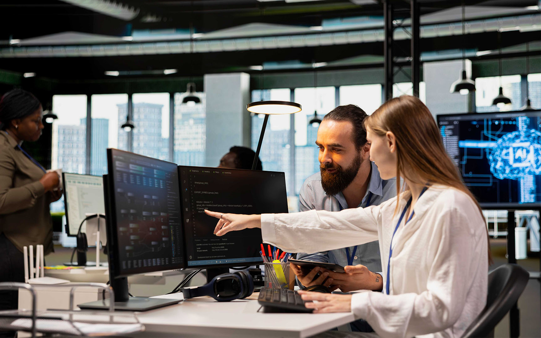 Two professionals collaborate at a workstation in a modern office, reviewing data displayed on multiple computer monitors.