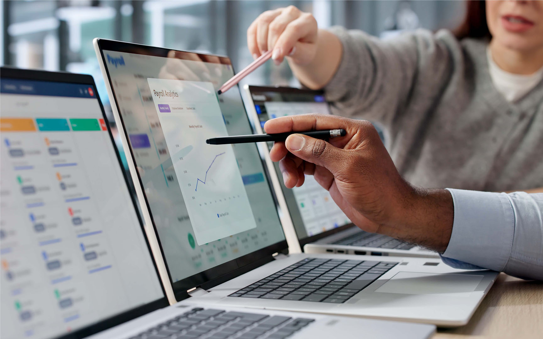 Close-up of business professionals reviewing payroll analytics on multiple laptop screens, pointing to charts and data visualizations during a collaborative discussion about workforce planning, reporting, and operational efficiency.