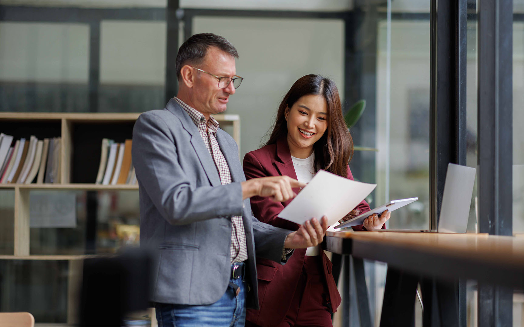 Two professionals standing in a modern office workspace reviewing printed documents together beside a workstation.