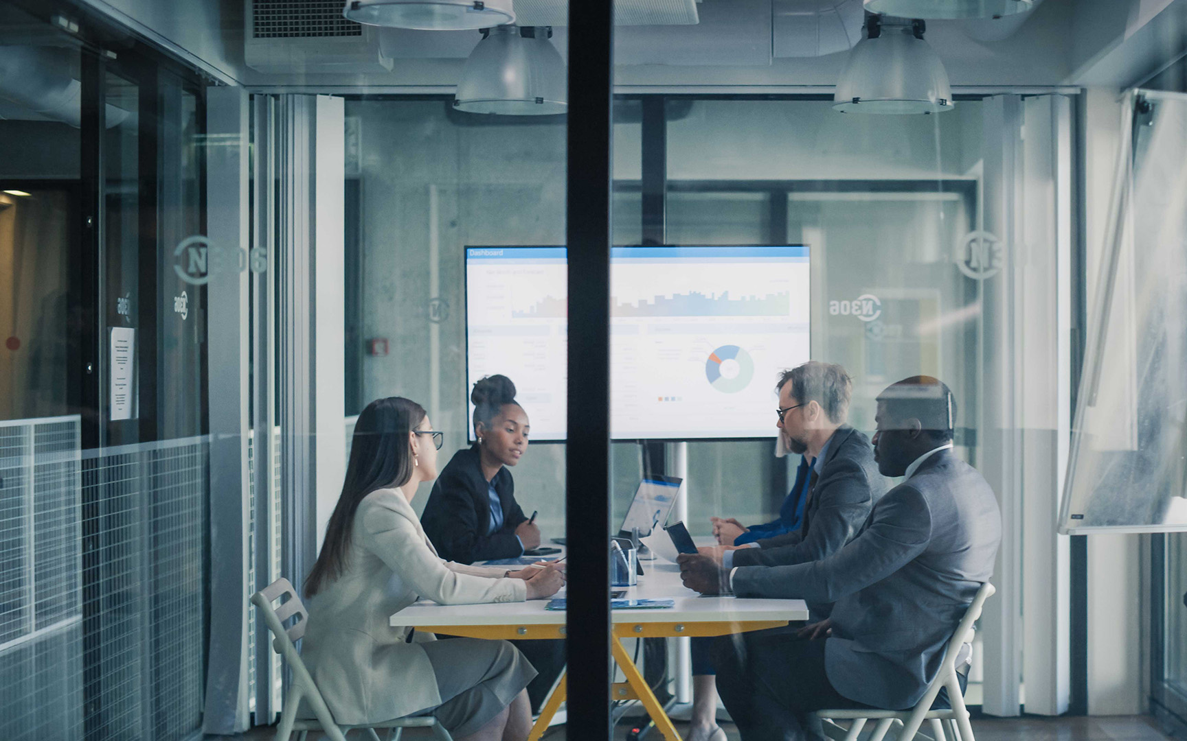 Professionals meeting in a modern glass-walled conference room, reviewing business performance dashboards and charts displayed on screens during a collaborative planning session.