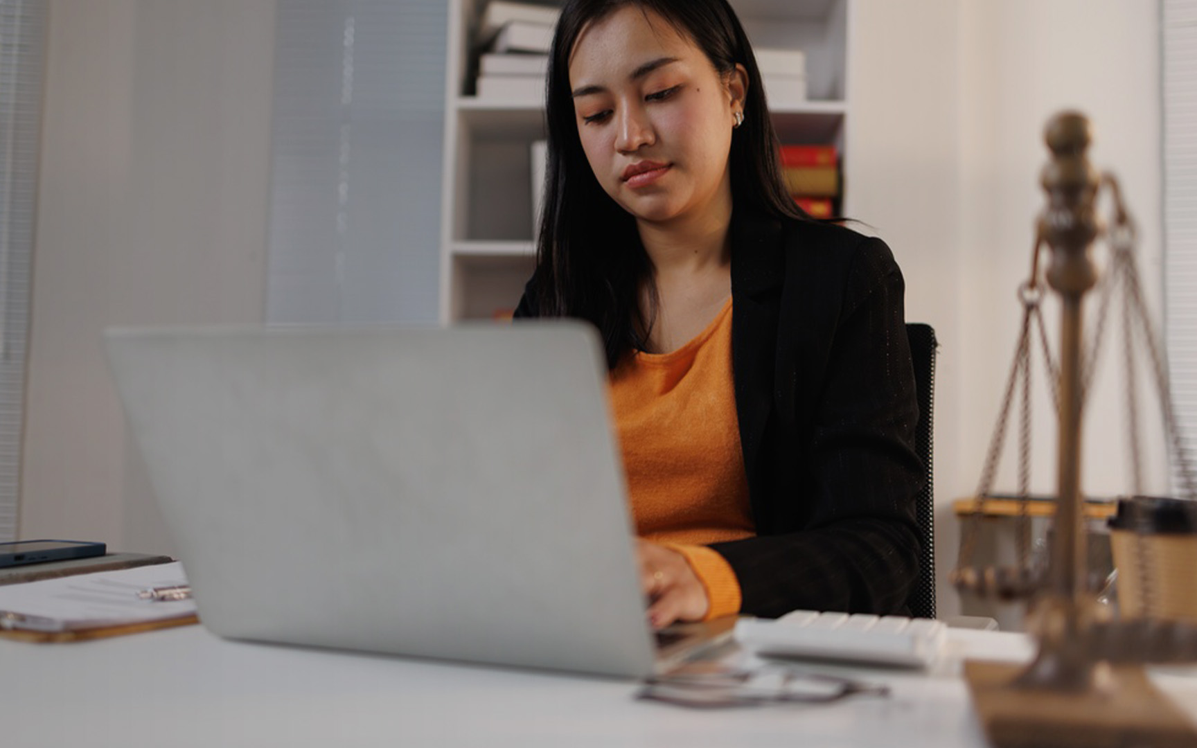 Person working on a laptop at a desk in an office setting, with a scales of justice ornament in the foreground, bookshelves in the background, and office accessories on the desk.