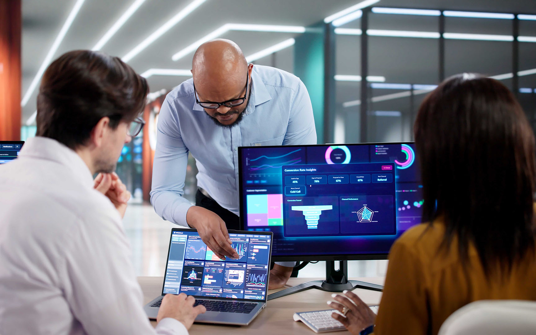 Three professionals collaborate in a modern office while reviewing business performance dashboards displayed on a laptop and external monitor.