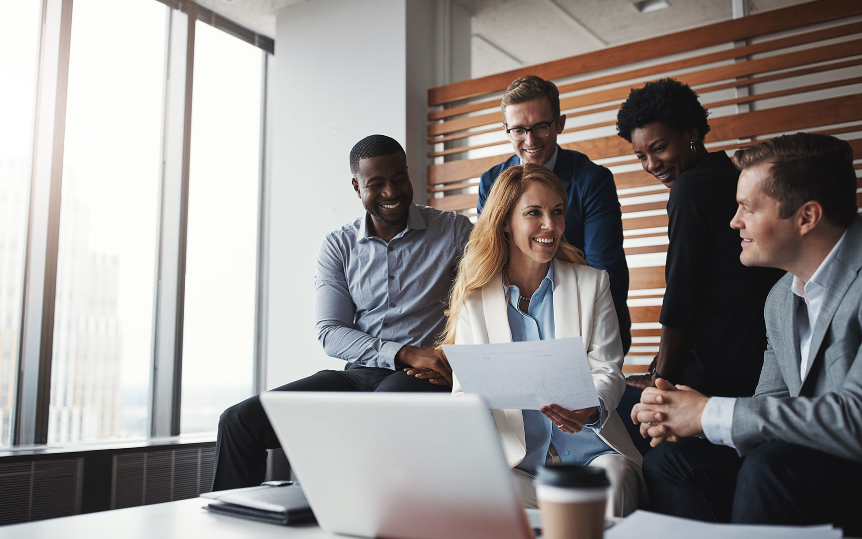 A group of business professionals gathered in a modern office meeting area, reviewing documents and collaborating around a laptop during a planning or strategy discussion.