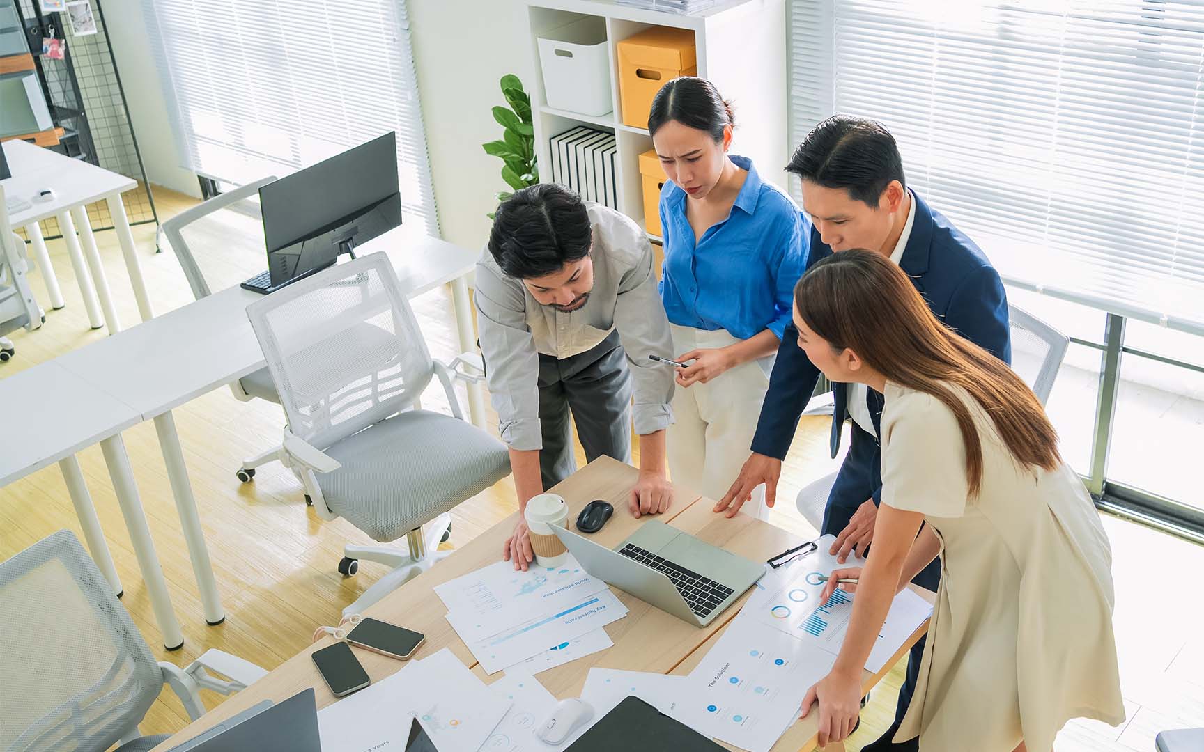 Four professionals gather around a desk in a bright office, reviewing charts and documents alongside a laptop during a collaborative planning and analysis session.