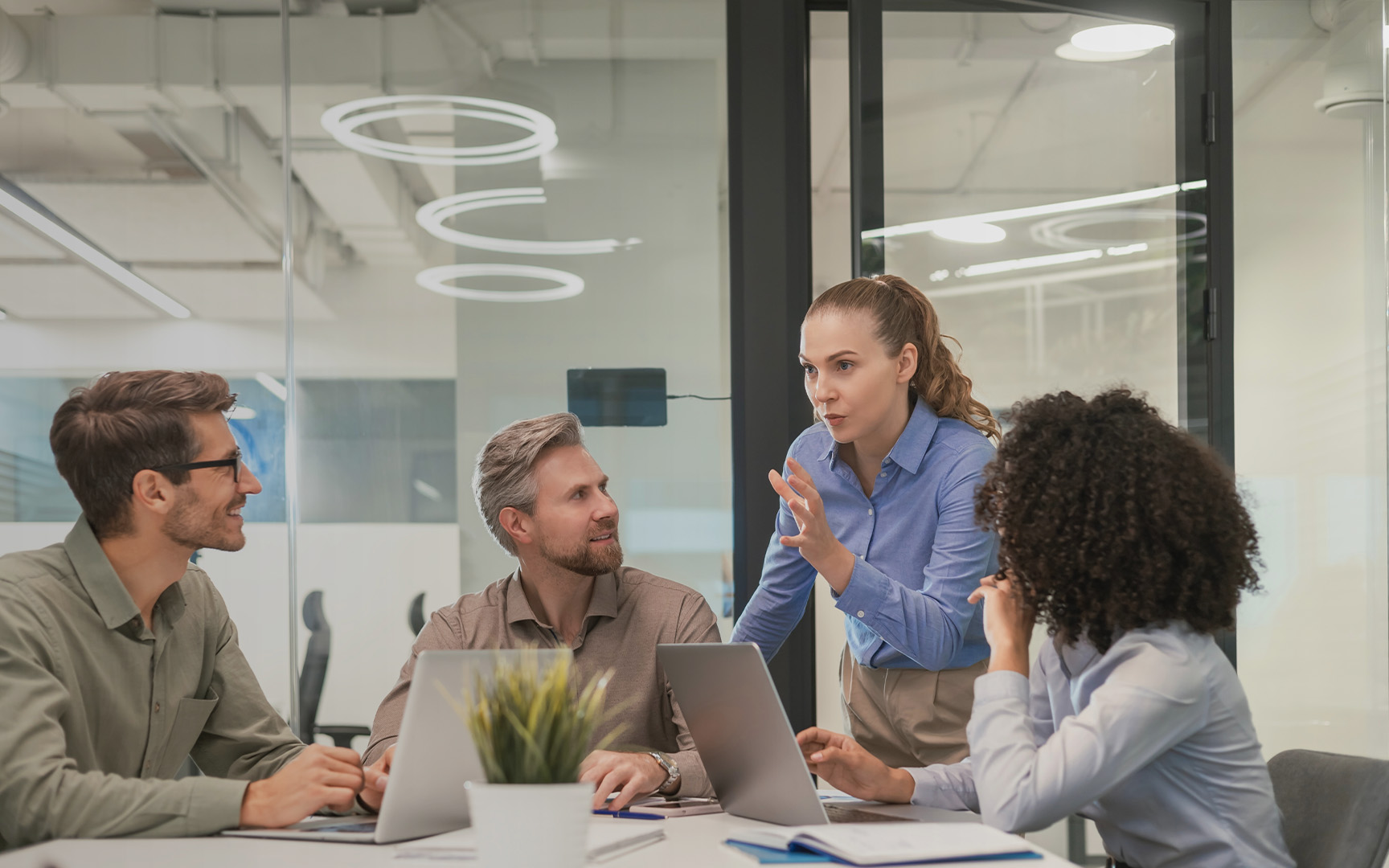 Four professionals collaborate around a conference table in a modern glass-walled office, using laptops while one person stands and presents ideas during a team discussion.