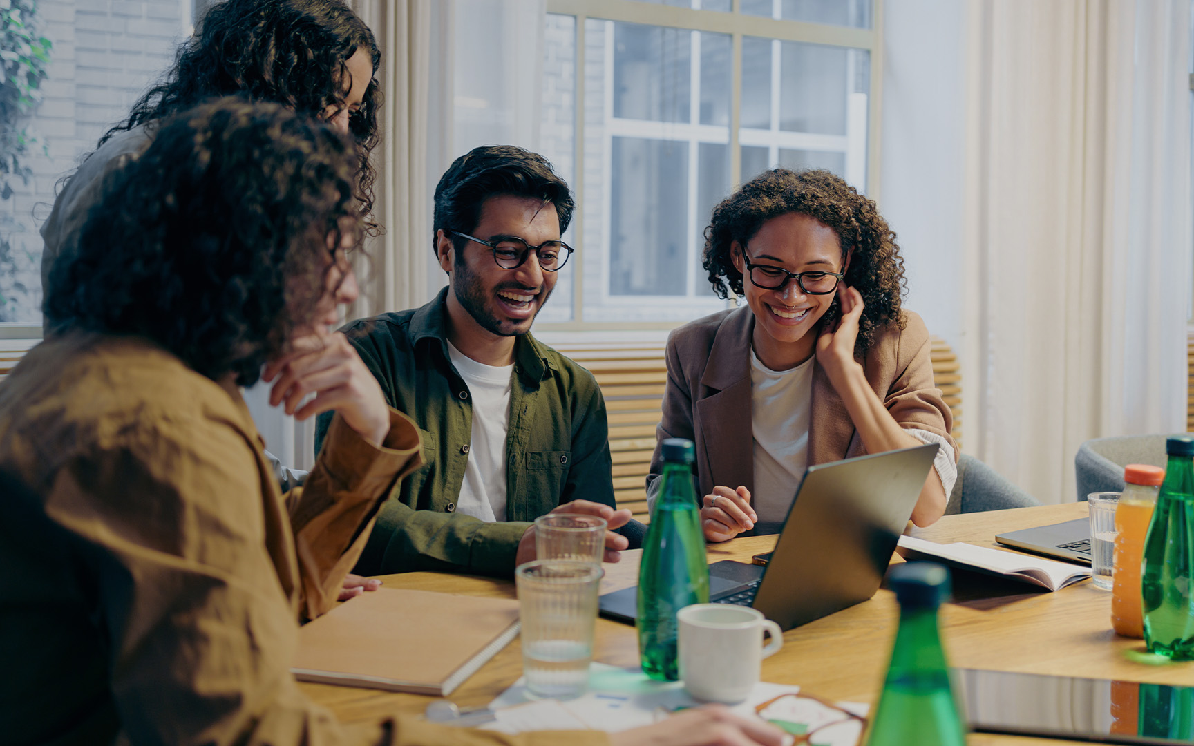 A group of professionals collaborate around a conference table in a bright office, reviewing information on laptops and discussing work.