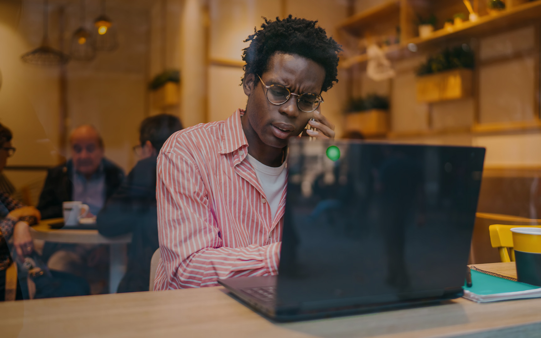 A professional works on a laptop while speaking on a phone in a café or coworking environment.