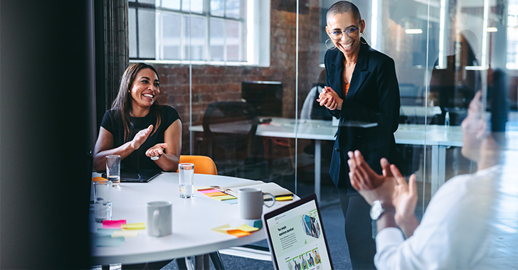 Three professionals discussing in a modern office setting with one person standing by a glass wall, while the other two are seated at a table with laptops and documents.