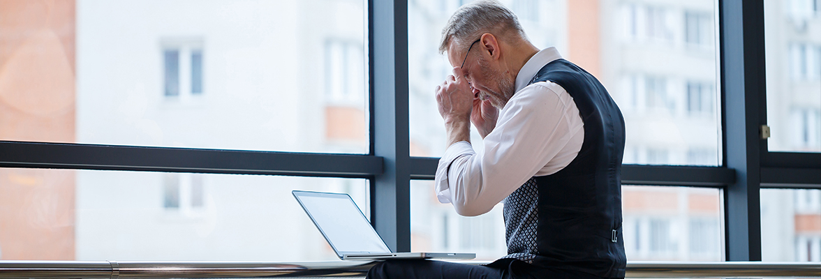 A person in business attire sits at a desk with a laptop, appearing stressed or thoughtful, in front of a large window with a cityscape view.