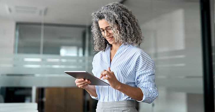 Person mit lockigem grauem Haar, Brille und gestreiftem Hemd, lächelt, während sie in einer modernen Büroumgebung ein Tablet verwendet.
