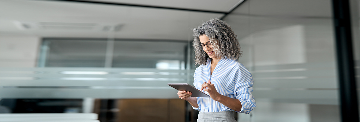 Person mit lockigem grauem Haar, Brille und gestreiftem Hemd, lächelt, während sie in einer modernen Büroumgebung ein Tablet verwendet.
