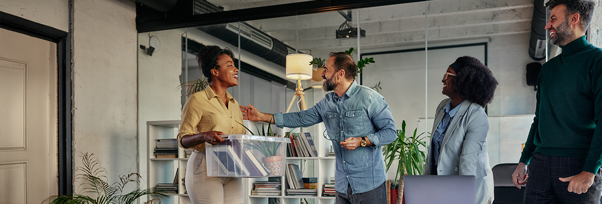 Two professionals shaking hands in an office setting with two other colleagues watching and smiling.