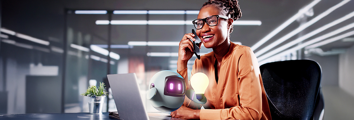 Henkilö istuu toimistossa työpöydän ääressä, puhuu puhelimeen ja käyttää kannettavaa tietokonetta, ja hänen vierellään on pieni robotti.