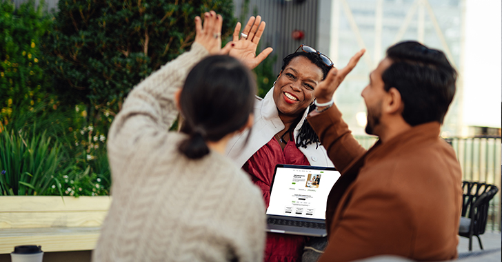 Three people engage in a cheerful conversation outdoors, with one person high-fiving another while the third person holds an open laptop.
