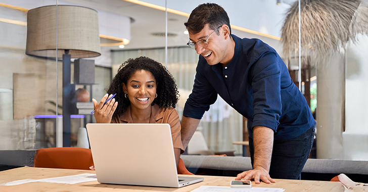 Two people are smiling and looking at a laptop in a modern office setting. They are sitting and standing at a table with papers. The environment is bright with a large window and contemporary decor.