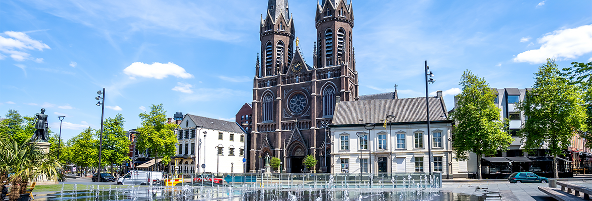 Stadsplein in Tilburg met fontein en de Heuvelse Kerk op de achtergrond, omringd door historische gebouwen en groen onder een blauwe lucht.