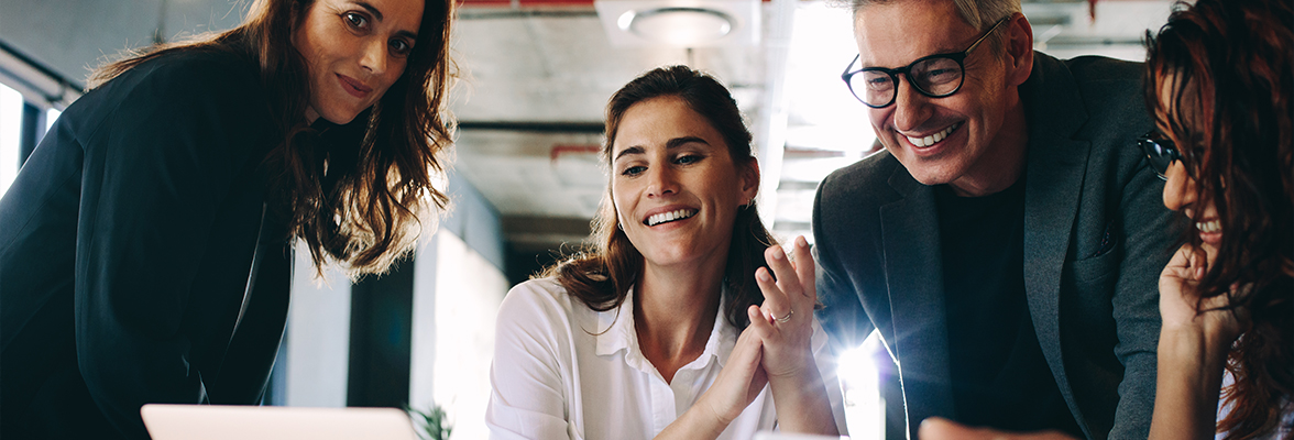 A group of people gathered around a laptop, appearing engaged and cheerful in a modern office setting. One person is holding a pen, and another is clapping. They are working collaboratively.
