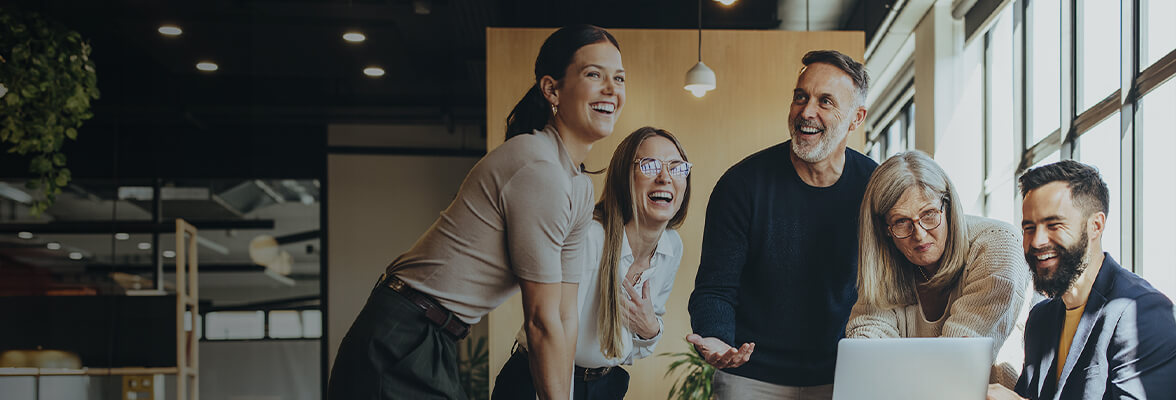 A group of people joyfully collaborating around a laptop in a modern office setting with large windows and indoor plants.