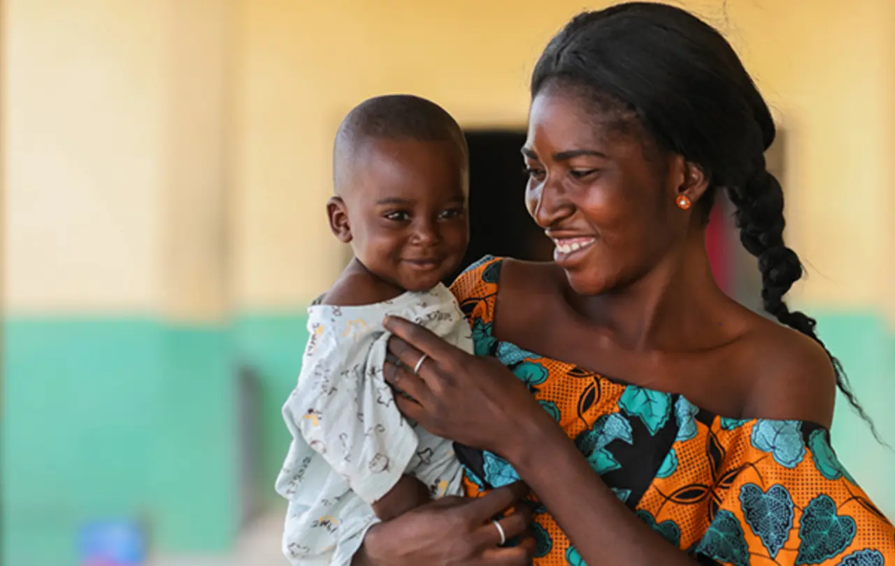 A smiling woman holds a baby, both with bright expressions, in a colorful setting.