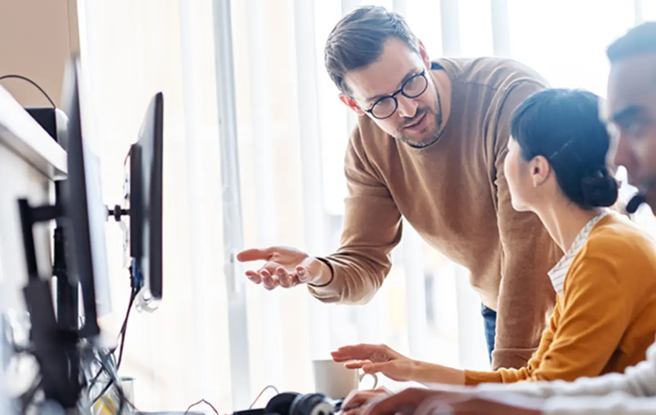 A person standing beside a seated colleague in an office setting, gesturing toward a computer screen while discussing work.