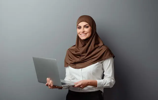 A woman wearing a brown hijab holds a laptop, smiling against a gray background. 